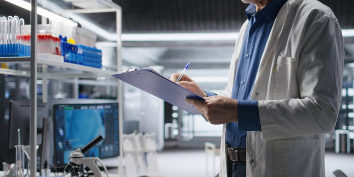 Laboratory technician in a lab coat writing on a clipboard beside scientific equipment and test tubes.